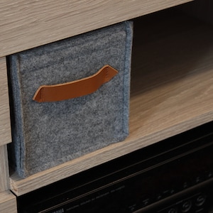 May include: A gray felt storage bin with a brown leather handle is placed on a wooden shelf. The bin is rectangular and appears to be part of a larger storage unit. The background includes a wooden cabinet and a black electronic device.