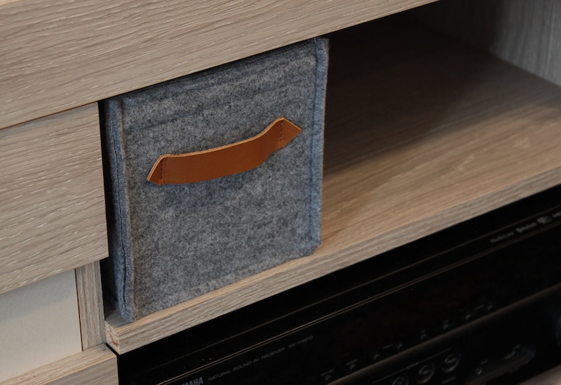May include: A gray felt storage bin with a brown leather handle is placed on a wooden shelf. The bin is rectangular and appears to be part of a larger storage unit. The background includes a wooden cabinet and a black electronic device.