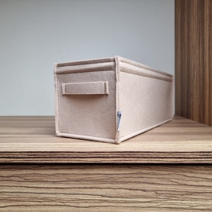 May include: A rectangular beige felt storage organizer with a handle on one side. The organizer is sitting on a wooden surface. The felt material appears to be soft and durable, suitable for organizing items.