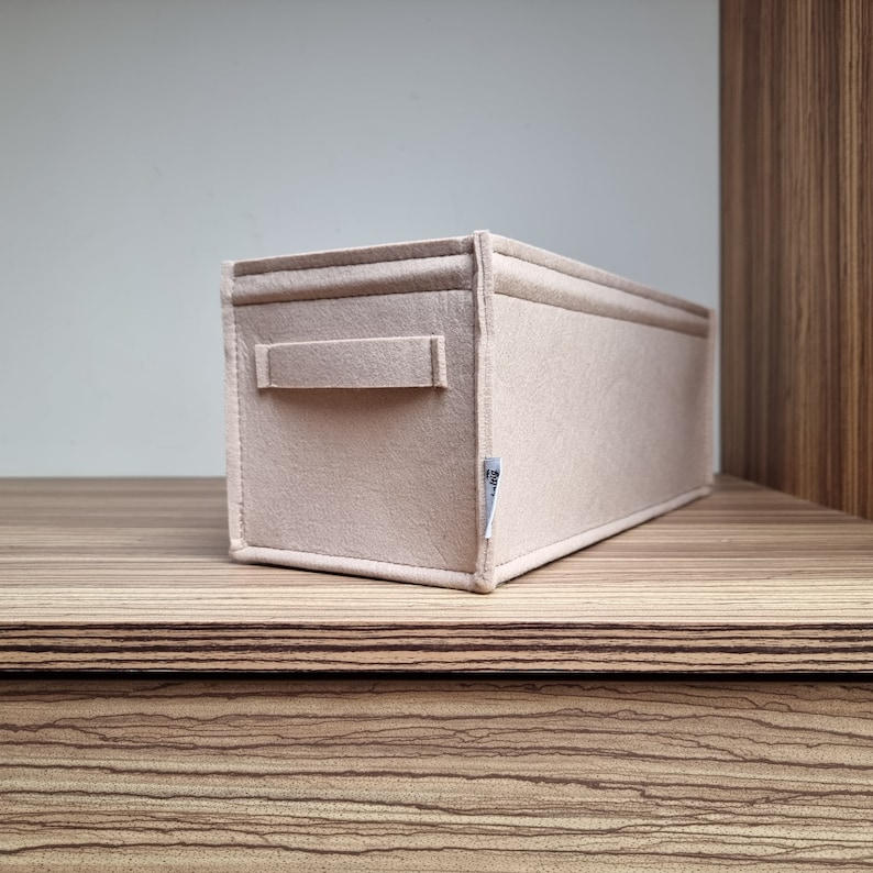 May include: A rectangular beige felt storage organizer with a handle on one side. The organizer is sitting on a wooden surface. The felt material appears to be soft and durable, suitable for organizing items.