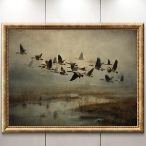 May include: A framed landscape painting depicts a flock of geese in flight over a misty marsh. The artwork features a gold-colored ornate frame. The geese are silhouetted against a muted, atmospheric background, with subtle reflections in the water.