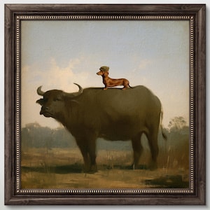 May include: A framed wall art print featuring a painting of a brown dachshund wearing a small green hat standing on the back of a large dark buffalo in a field. The artwork is enclosed in a dark brown wooden frame with a decorative beaded inner edge.