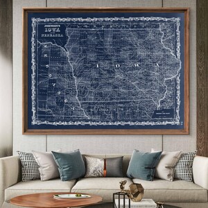 May include: A vintage map of Iowa and Nebraska printed in white on a dark blue background. The map is framed in a dark wood frame and is hanging on a wall in a living room setting.