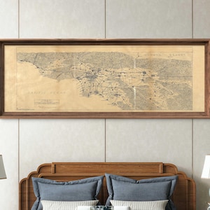 May include: A framed vintage map of Los Angeles, California, with a dark wood frame. The map is printed on aged paper and shows the city's layout, including the Pacific Ocean and surrounding areas. The map is displayed above a bed with blue pillows and a wooden headboard.