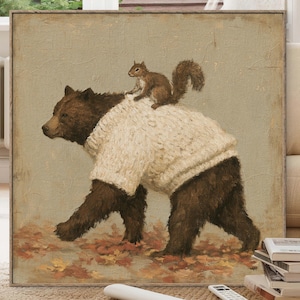 May include: A square framed painting depicting a brown bear wearing a thick, cream-colored cable-knit sweater. A small brown squirrel is perched on the bear's back. The bear is walking across a ground covered in scattered orange and red autumn leaves.