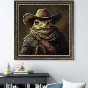 Op de afbeelding: Een schilderij van een kikker die een cowboyhoed en een bruine sjaal draagt. De kikker kijkt recht naar de kijker. Het schilderij is ingelijst in een donkerbruin lijst.