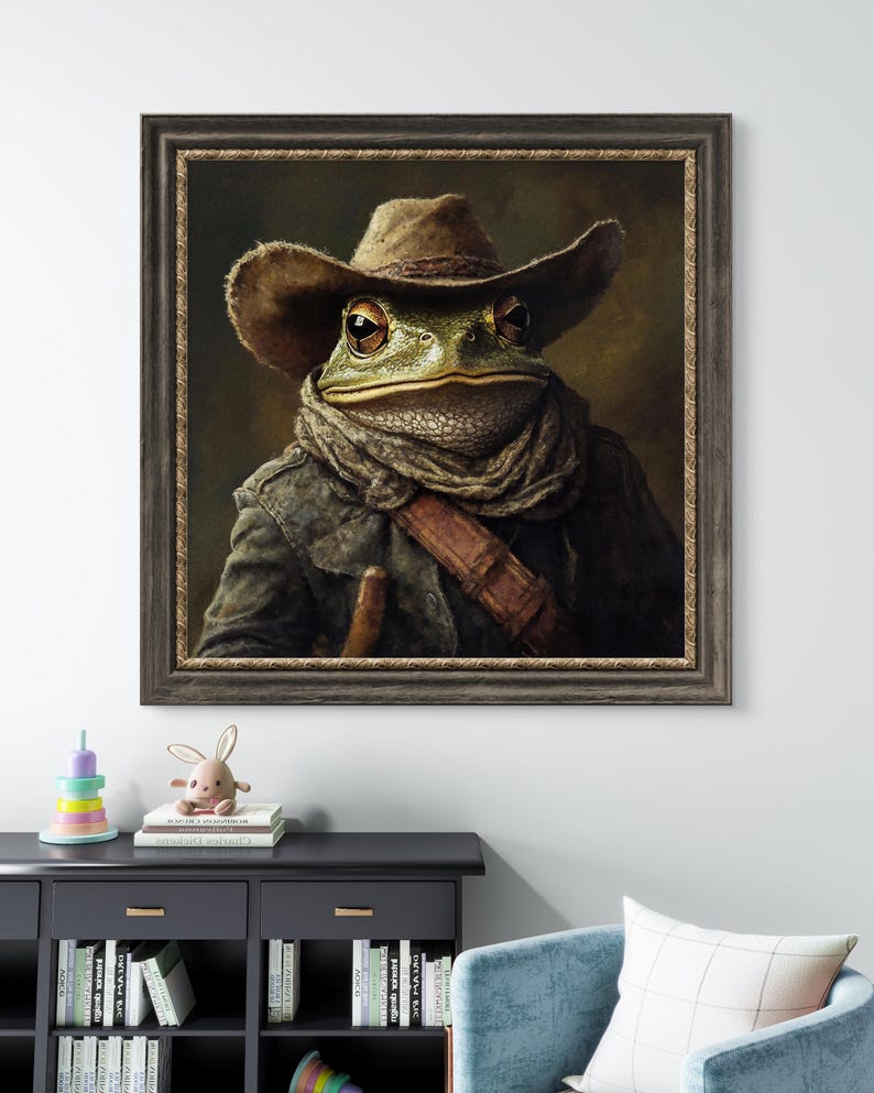 Op de afbeelding: Een schilderij van een kikker die een cowboyhoed en een bruine sjaal draagt. De kikker kijkt recht naar de kijker. Het schilderij is ingelijst in een donkerbruin lijst.