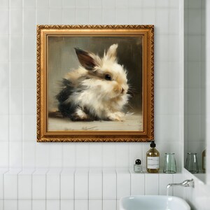 Puede incluir: Pintura enmarcada de un conejito esponjoso con pelaje negro, blanco y marr&oacute;n. La obra de arte est&aacute; en un marco dorado adornado y cuelga de una pared de azulejos blancos sobre un lavabo blanco. La pintura es de estilo suave y realista.