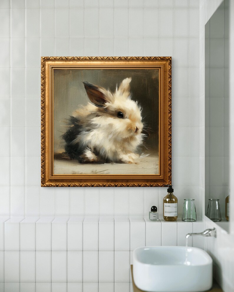 Puede incluir: Pintura enmarcada de un conejito esponjoso con pelaje negro, blanco y marr&oacute;n. La obra de arte est&aacute; en un marco dorado adornado y cuelga de una pared de azulejos blancos sobre un lavabo blanco. La pintura es de estilo suave y realista.