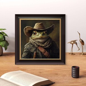 Op de afbeelding: Een ingelijst schilderij van een groene pad die een bruine cowboyhoed en een bruine sjaal draagt. De pad kijkt de kijker recht aan met een serieuze uitdrukking. Het schilderij is geplaatst tegen een donkerbruine achtergrond.