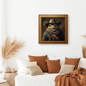 Op de afbeelding: Een ingelijst portret van een kikker die een cowboyhoed en een sjaal draagt. De kikker kijkt rechtstreeks naar de kijker. De lijst is goudkleurig en versierd.