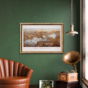 Puede incluir: Una ilustraci&oacute;n de estilo vintage del lago Chautauqua, Nueva York, enmarcada en un marco dorado y colgada en una pared verde. Un sill&oacute;n de cuero marr&oacute;n se encuentra frente a la obra de arte, y un tocadiscos vintage con un cuerno dorado se encuentra en un soporte de madera a la derecha.