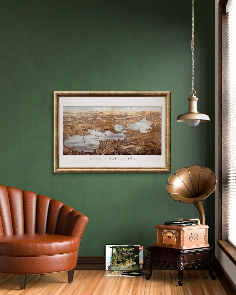 Puede incluir: Una ilustraci&oacute;n de estilo vintage del lago Chautauqua, Nueva York, enmarcada en un marco dorado y colgada en una pared verde. Un sill&oacute;n de cuero marr&oacute;n se encuentra frente a la obra de arte, y un tocadiscos vintage con un cuerno dorado se encuentra en un soporte de madera a la derecha.