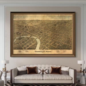 May include: An antique map of Columbus, Ohio, printed in 1869. The map shows the city's streets, buildings, and river. The map is framed in a brown wood frame and is hanging on a wall in a living room.