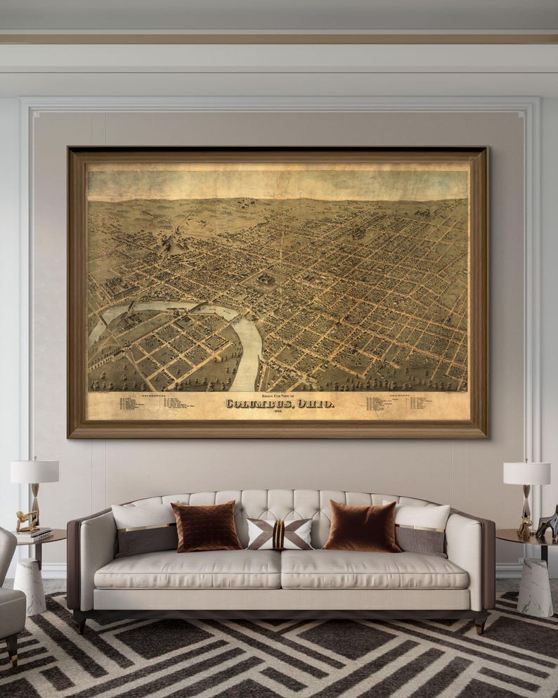 May include: An antique map of Columbus, Ohio, printed in 1869. The map shows the city's streets, buildings, and river. The map is framed in a brown wood frame and is hanging on a wall in a living room.