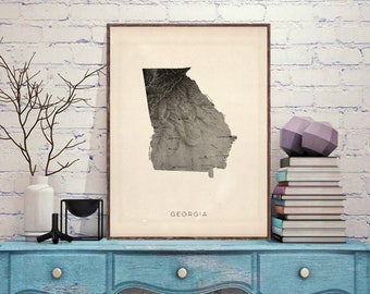 May include: A framed grayscale topographic map of Georgia, with the state's name printed below. The artwork is displayed on a blue table with decorative items, including a vase with branches, a black candle holder, and a stack of books.