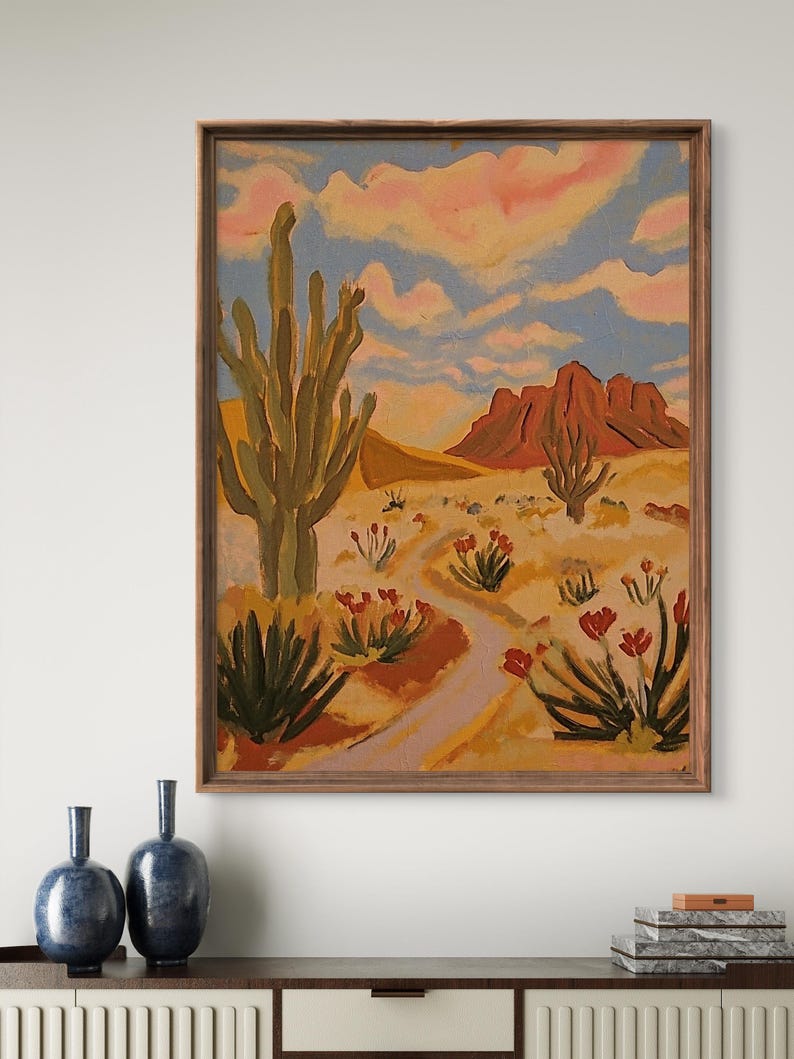 May include: A painting of a desert landscape with a tall cactus, red mountains, and a winding path. The sky is a light blue with pink clouds. The painting is framed in a brown wood frame.