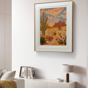 May include: A framed painting of a desert landscape with a tall cactus, red flowers, and a winding path leading to a mountain range in the distance. The sky is a vibrant blue with pink and orange clouds.