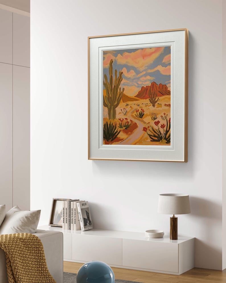 May include: A framed painting of a desert landscape with a tall cactus, red flowers, and a winding path leading to a mountain range in the distance. The sky is a vibrant blue with pink and orange clouds.