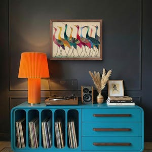 May include: A teal blue cabinet with a built-in record player, orange lamp, and framed artwork of colorful birds. The cabinet has open slots for vinyl records and drawers with wooden handles. A small speaker and decorative vase are also on top.