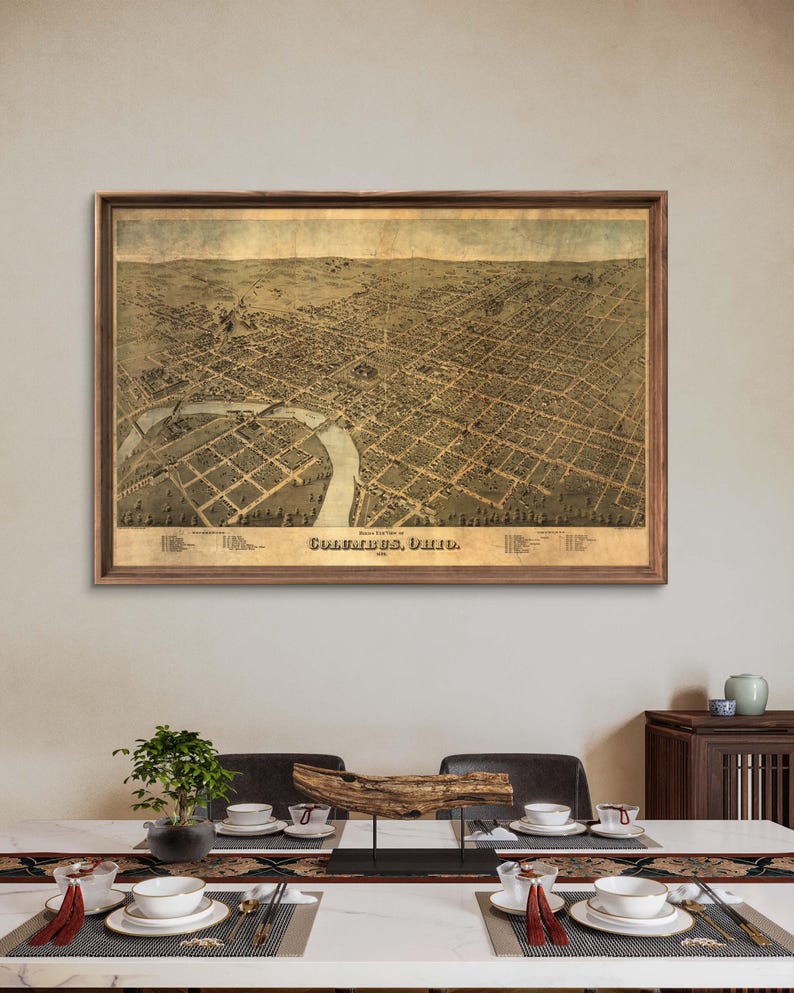 May include: An antique map of Columbus, Ohio, framed in a brown wood frame. The map is printed on aged paper and shows the city's streets and buildings in detail. The map is hanging on a wall in a room with a dining table and chairs.