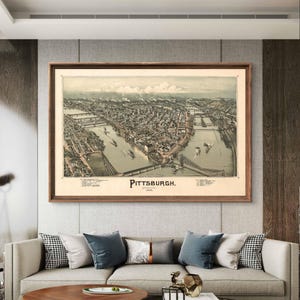 May include: A vintage map of Pittsburgh, Pennsylvania, in a brown wooden frame, hanging on a gray wall above a beige sofa with pillows. The map shows the city's layout with rivers and bridges.