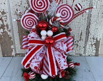 May include: A festive Christmas centerpiece featuring red and white swirl lollipops, candy canes, and ornaments. The arrangement includes a large red and white striped bow, set on a bed of green pine needles, creating a holiday theme.
