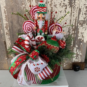 May include: A festive Christmas centerpiece featuring a red and green glittered container filled with greenery, candy canes, and a nutcracker figurine. The arrangement includes a large bow with red and white striped ribbon, and a ballerina figurine.