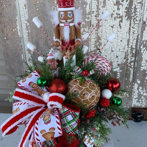 May include: A festive Christmas centerpiece featuring a wooden nutcracker with a red hat and white beard. The arrangement includes gingerbread-themed ornaments, red and green baubles, and a large bow with gingerbread man patterns.