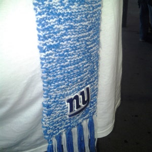 May include: A blue and white knitted scarf with fringe. The scarf features the letters "NY" in a navy blue emblem. The scarf is draped over a white t-shirt.