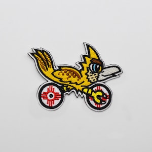 May include: A yellow embroidered patch of a cartoon roadrunner riding a motorcycle with red and white wheels. The wheels have a stylized sun symbol in the center.