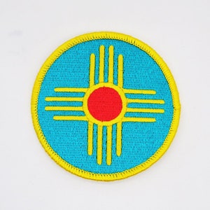 May include: An embroidered patch featuring the Zia symbol, the state symbol of New Mexico, in yellow and red on a blue background.