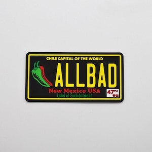 Pode incluir: Uma placa de matrícula preta e amarela com o texto "ALLBAD" em letras amarelas. A placa também tem o texto "CHILE CAPITAL OF THE WORLD", "New Mexico USA", "Land of Enchantment", e "47th 1912". Uma pimenta verde e vermelha está no lado esquerdo da placa.