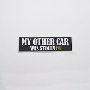 以下が含まれることがあります： 白黒のステッカーに、白い文字で「My other car was stolen NM」と書いてあります。「NM」は黄色の正方形の中にあります。