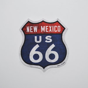 Puede incluir: Un cartel de estilo vintage azul y rojo de la Ruta 66 con el texto "New Mexico" y "US 66" en blanco. El cartel tiene un aspecto desgastado y envejecido.