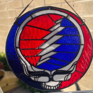 May include: Round stained glass art piece featuring a skull with a lightning bolt design. The artwork uses red, white, and blue glass, with a black border. The piece is suspended by a thin, natural-colored cord.