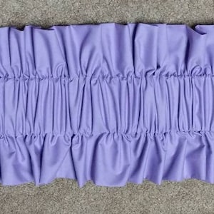 May include: A close-up of a lavender fabric ruffle trim. The trim is gathered and has a soft, flowing texture.