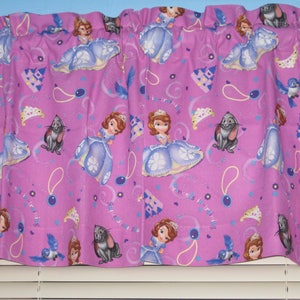 May include: Pink fabric valance with a princess theme. The valance features a repeating pattern of a princess in a blue dress, a castle, a crown, a rabbit, and a bluebird. The text "Princess Sofia" is repeated throughout the valance.