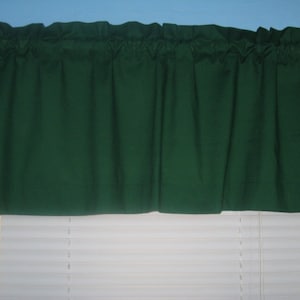 May include: A dark green valance with a ruffled edge hangs above a white window blind.