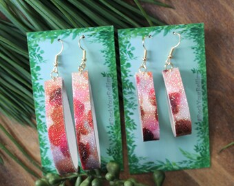 May include: A pair of dangle earrings with a pink and white glitter pattern on a light blue background. The earrings are on a card with the text "#RockYourPellums".