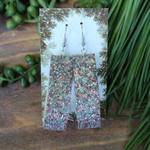 May include: A pair of dangle earrings with silver hooks and a rectangular shaped charm. The charm is made of a glittery material with a mix of pink, blue, and green glitter.