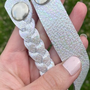 May include: Two white iridescent leather wrist cuffs with silver snap closures. One cuff is a flat strap, and the other is a braided design. The cuffs have a textured, shimmering appearance, reflecting various colors.
