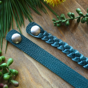 May include: Two teal leather bracelets with silver metal clasps. One bracelet is braided and the other is flat.