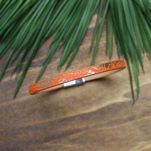 May include: A thin orange leather bracelet with a silver magnetic clasp. The bracelet has a textured pattern that resembles scales.