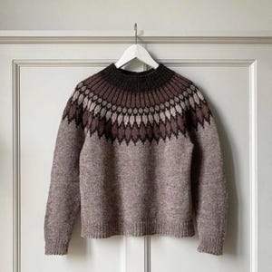 May include: A long-sleeved knit sweater featuring a dark brown ribbed crew neckline and a yoke decorated with a geometric pattern in shades of dark brown, light brown, and cream. The body and sleeves are a solid heathered light brown knit.