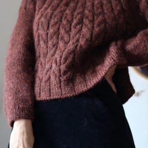 May include: A rust-colored, long-sleeved sweater featuring a vertical cable knit pattern and a ribbed hem. The garment is crafted from a textured, fuzzy yarn and is paired with a solid black skirt. The sweater has a high neckline and long sleeves.