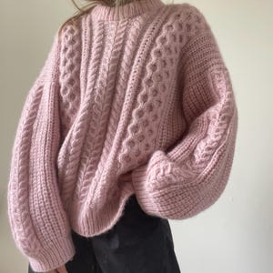 Könnte beinhalten: Ein hellrosa Langarmpullover mit einem groben Zopfstrickmuster auf der Vorderseite und den Ärmeln, einem gerippten Rundhalsausschnitt und gerippten Bündchen. Das Kleidungsstück hat eine lockere, übergroße Passform mit einer strukturierten Optik.