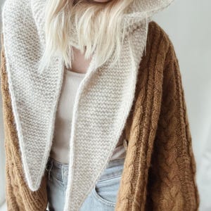 Peut inclure: Une écharpe triangulaire en tricot couleur crème avec une texture au point mousse, drapée sur un gilet en tricot torsadé marron. Le gilet présente un motif torsadé épais et texturé, porté sur un débardeur côtelé clair et un jean.