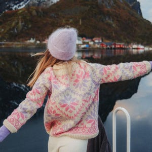 May include: A long-sleeve sweater featuring a cream base with a repeating pink and lavender snowflake pattern, paired with a matching lavender knit beanie and lavender gloves. The sweater has a ribbed crew neckline, cuffs, and hem.
