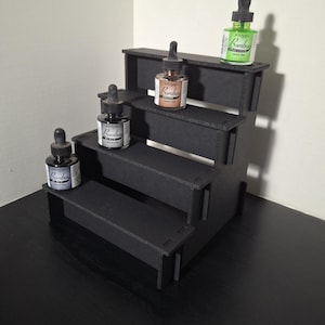 May include: A black, tiered display stand holding several bottles of ink. The ink bottles are various colors, including brown, green, and blue. The stand is made of black material and has four levels, each holding a bottle of ink.